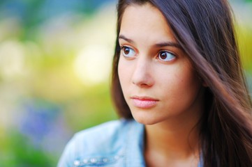 Obraz premium Portrait of young long haired cute young brunette woman with sad brown eyes on a summer day on blurred colorful background, outside, close-up