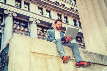 American Businessman with beard, mustache traveling, working in New York, wearing cadet blue suit,  white undershirt, brown leather shoes, sitting on street, reading, working on laptop computer..