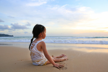 Fototapeta na wymiar Rear view of asian girl sitting and playing sand on the beach at the sunset.