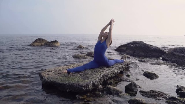 Young woman in bodysuit practicing yoga on the beach above sea at amazing sunrise. Fitness, sport, yoga and healthy lifestyle concept. Girl doing complicated yoga asana