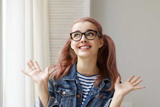 Attractive fascinated young woman shopaholic wearing trendy denim jacket and eyeglasses gesturing with open palms and smiling broadly, feeling excited about high sale prices in clothing stores