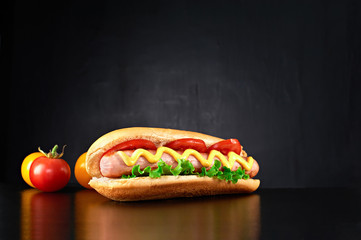 Big hotdog with sausage tomatoes, mustard, ketchup and salad isolated on black background. Front...