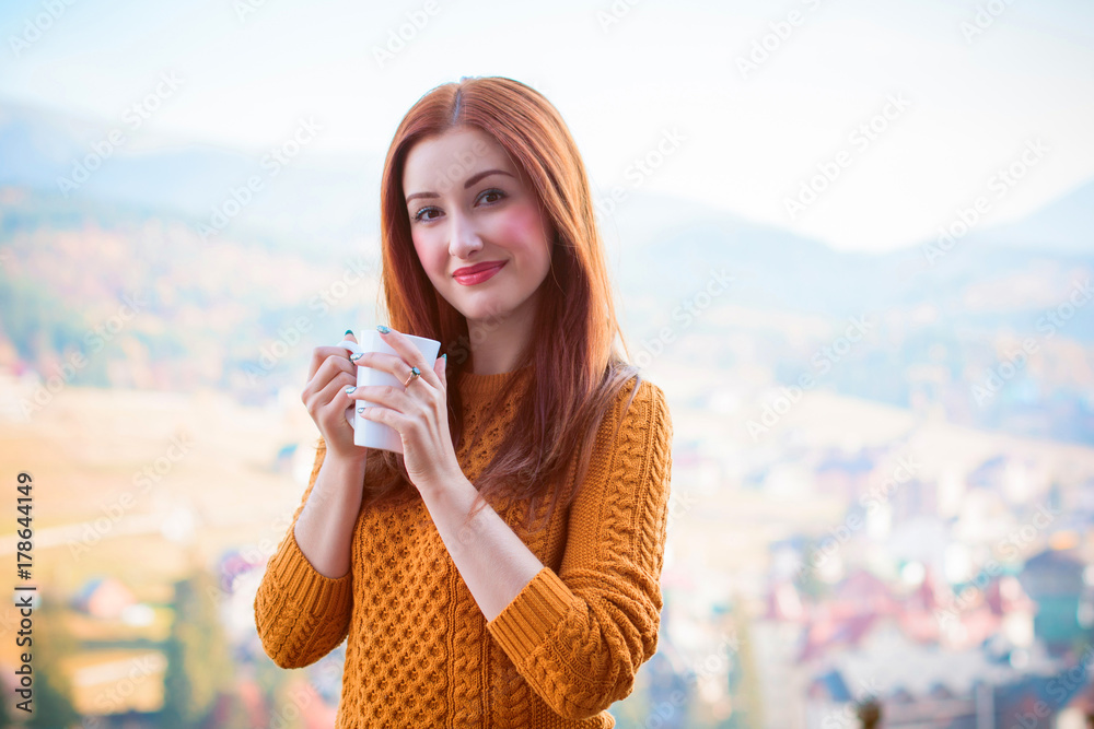 Wall mural Cute and nice young woman drinking coffee at terrace on a sunny morning in mountain. Pretty lady in stylish casual yellow sweater and shorts. Warm soft cozy image, breakfast on the balcony  