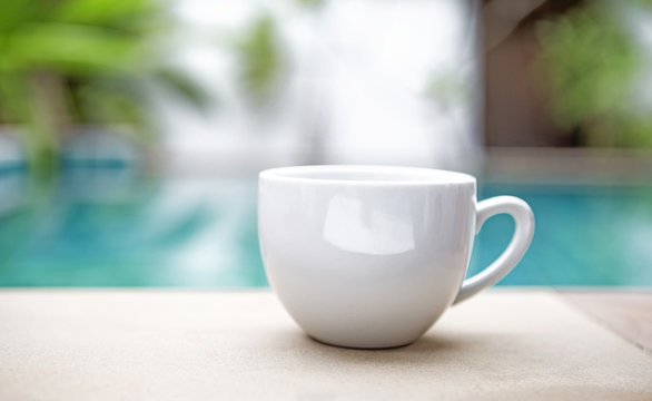 White ceramic coffee cup over blurred swimming pool, outdoor day light