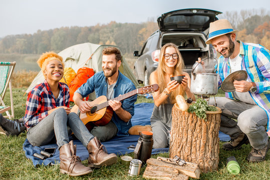 Multi ethnic group of friends dressed casually having a picnic, cooking soup with cauldron, playing guitar during the outdoor recreation near the lake