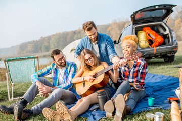 Multi ethnic group of friends dressed casually having fun playing guitar during the outdoor recreation with tent, car and hiking equipment near the lake