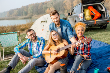 Obraz na płótnie Canvas Multi ethnic group of friends dressed casually having fun playing guitar during the outdoor recreation with tent, car and hiking equipment near the lake