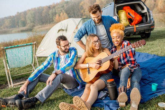 Multi Ethnic Group Of Friends Dressed Casually Having Fun Playing Guitar During The Outdoor Recreation With Tent, Car And Hiking Equipment Near The Lake