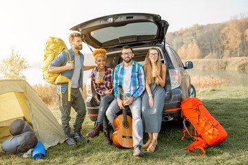Multi ethnic group of friends sitting at the car trank during the outdoor recreation with tent and...