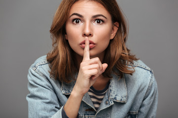 Naklejka premium Close-up portrait of beautiful brunette woman in jeans jacket showing silence gesture, looking at camera