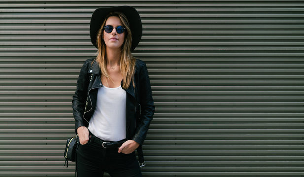 Pretty blonde hipster girl wearing white blank t-shirt leather jacket and sunglasses is looking at camera while standing beside the gray wall.Beautiful woman wearing hat is standing on wall background