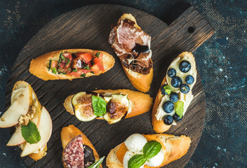 Italian crostini with various toppings on round wooden serving board over black plywood background, top view