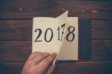 New Year 2018 is coming concept. Hand flips notepad sheet on wooden table. 2017 is turning, 2018 is...