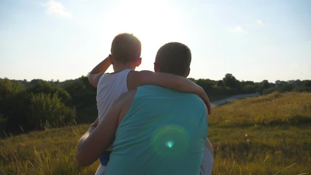 Father with his little son spending time together outdoor at sunset. Dad and child sitting at green grass on the hill and looking to the nature around them. Slow motion Rear back view