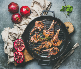 Barbecue dinner. Grilled lamb meat chops with onion and rosemary in black cast iron pan served with fresh pomegranates on wooden board over grey concrete background, top view. Slow food concept © sonyakamoz