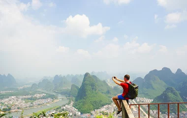 Rollo Hiker with backpack take a picture on his smartphone from top of the hill with beautiful view on Yangshuo city with mountains around. Yangshuo, China © upslim