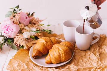 Delicious healthy breakfast with two fresh flaky croissants served on a plate with two cups of coffee and a bouquet of summer wildflowers on kitchen table. Woman's hand pouring coffee