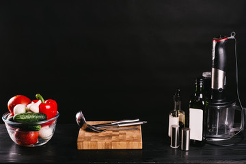 Ingredients for cooking gazpacho,Spanish cold soup made from ripe tomatoes and fresh cucumbers. Glass bowl of vegetables, wooden cutting board,bottle of olive oil,vinegar,blender on a black table