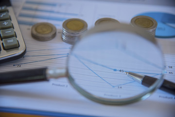 Business accessories (notebook, magnifier, calculator, planchette, tablet, fountain pen, notebook, glasses) and graphics, tables, charts on a wooden office desk. Soft focus