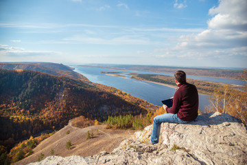 Naklejka na ściany i meble a man sits on top of a mountain, admires the scenery and works on a tablet