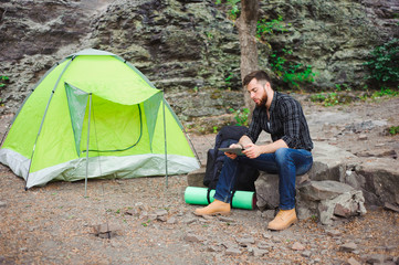 Traveler near a tent with a tablet in the evening. Rocky mountains background.