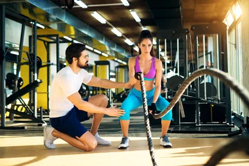 Tuinposter Fitness woman in training doing exercises with battle rope in the gym with a trainer next to her. © dusanpetkovic1
