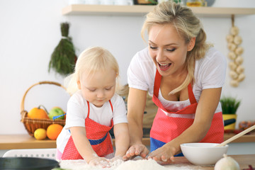 Little girl and her blonde mom in red aprons  playing and laughing while kneading the dough in the kitchen. Homemade pastry for bread, pizza or bake cookies