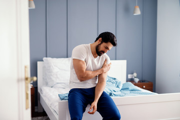 Attractive strong man sitting on the bed and holding painful shoulder with another hand.