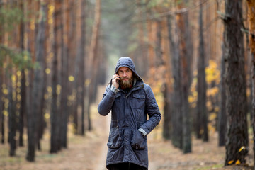 A man in a gray jacket stands on the road in the middle of a pine forest and talks on the phone 