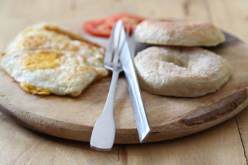 Breakfast; donut, fried egg and tomato with knife and fork on wooden plate.