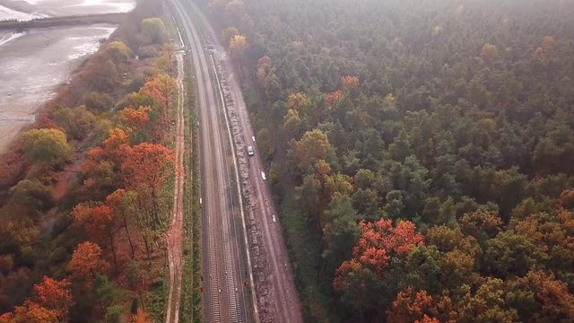 Aerial Shot Of A Car Coming Into The Scene From The Top. The Camera Moves To Track And Follow The Car As It Exits Through The Bottom Of The Scene, Which Is A Road Among Trees, Next To A Railway.