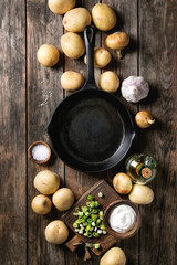 Ingredients for cooking dinner. Raw whole washed organic potatoes, onion, garlic, salt, olive oil, cast-iron pan, spring onion and cream-fresh over old wooden plank background. Top view with space