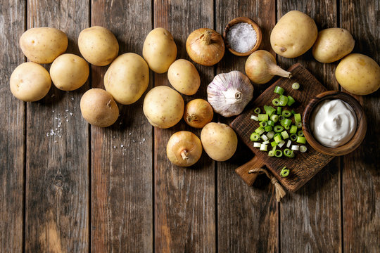 Ingredients for cooking dinner. Raw whole washed organic potatoes, onion, garlic, salt, spring onion and cream-fresh on cutting board over old wooden plank background. Top view with space