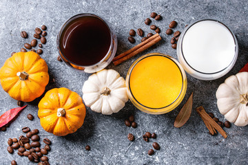 Ingredients for cook spicy pumpkin latte. Glasses with black coffee, pumpkin milk, cream with spices, coffee beans and decorative pumpkins above over gray texture background. Top view, space.