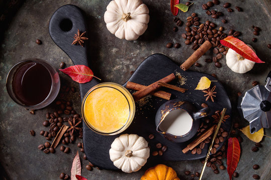 Ingredients for cook spicy pumpkin latte. Glasses with black coffee, pumpkin milk, jug of cream on black serving board. Spices, coffee beans, autumn leaves above over dark background. Top view, space.