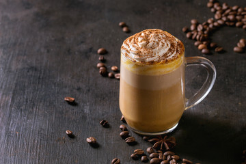 Glass of spicy pumpkin latte with whipped cream and cinnamon standing on black serving board. Coffee beans and spices above. Dark background.