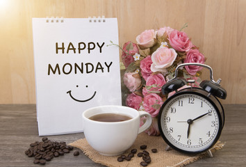 Hot cup of coffee and alarm clock on wood table with rose flower and happy monday note on morning sunlight.