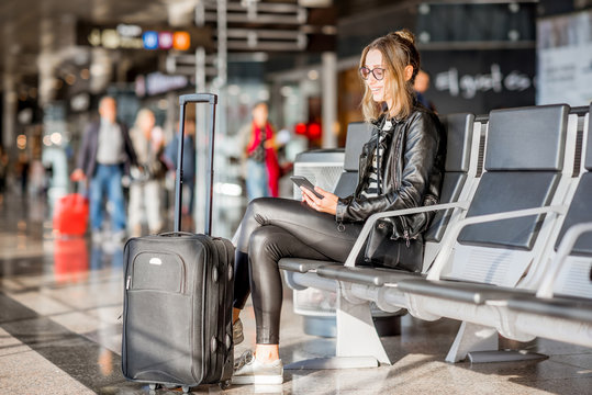 Young business woman in leather jacket and pants sitting with phone and suitcase at the departure hall of the airport waiting for the flight