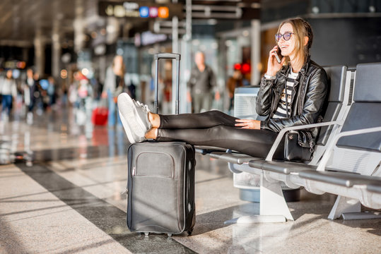 Young business woman in leather jacket and pants sitting with phone and suitcase at the departure hall of the airport waiting for the flight