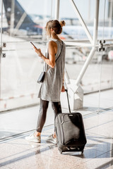 Young woman standing with phone near the airport window waiting for the flight