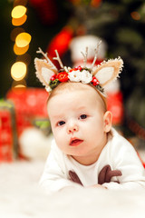 Charming little baby with deer ears lies on the carpet before a Christmas tree