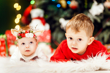 Charming little baby with deer ears and her little brother lie on the carpet before a Christmas tree