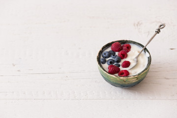 Top view on small bowl with organic yogurt with blueberries and raspberries on white wooden background. Girl having healthy breakfast at home. Healthy eating, snack.