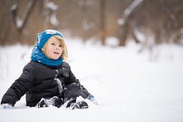 Fototapeta na wymiar Portrait of cute kid boy in warm clothes and hat sitting on the snow and playing with it in the park. Child having fun outdoors. Winter, lifestyle concept.