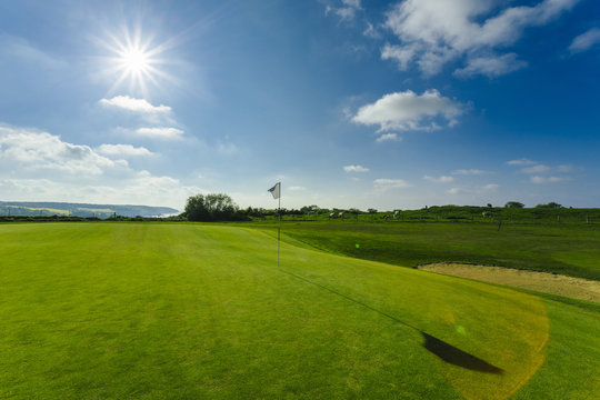 View of a green golf course, hole and flag on a bright sunny day. Sport, relax, recreation and leisure concept. Summer landscape with sunbeams