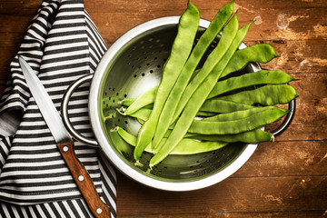 Chauchas y judías verdes crudas dentro de un colador de aluminio junto a un cuchillo y un paño de...