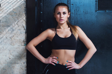 Confident Caucasian fitness model in black sportswear standing hands on hips looking at camera posing against black door