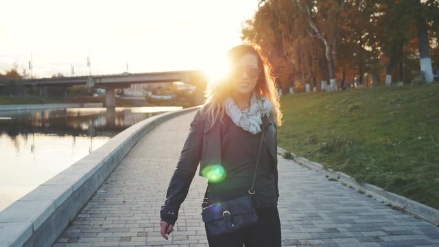 Attractive woman in mirrored sunglasses, a black leather jacket, black jeans walking along the river Bank in the city, turns to camera and smiles in front the setting sun. Waving hair. city,bridge.