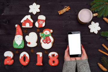 Mobile smart phone with blank screen for app presentation in woman hands, copy space. Christmas background with gingerbread cookies and cup of cocoa. Flat lay, top view. Application mock up template