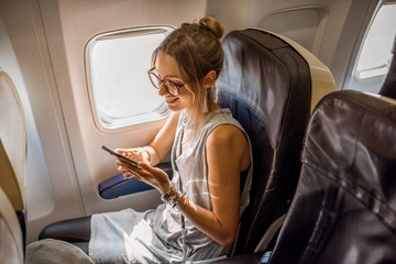 Young woman sitting with phone on the aircraft seat near the window during the flight in the airplane